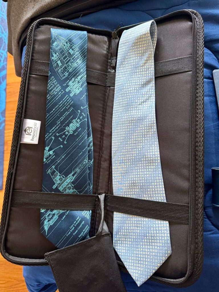An open, black, zip-around travel tie case with two neckties secured by elastic straps. The tie on the left is dark blue with a detailed, light-cyan-line-art schematic of an X-wing fighter from Star Wars, showing internal components with technical callouts. The tie on the right is light grey with a dense, repeating, multi-colored (blue and gold) pattern of mixed-case letters and numbers. This text represents a specific block of Enigma ciphertext, referencing Bletchley Park.
